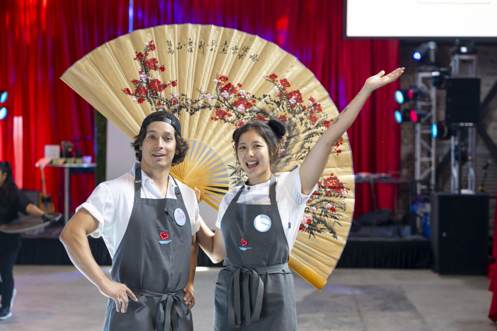 Chefs Sarah Cai with her right arm in the air and Chef Arturo Leighton smiling for the camera in front of a large foldable paper fan decoration