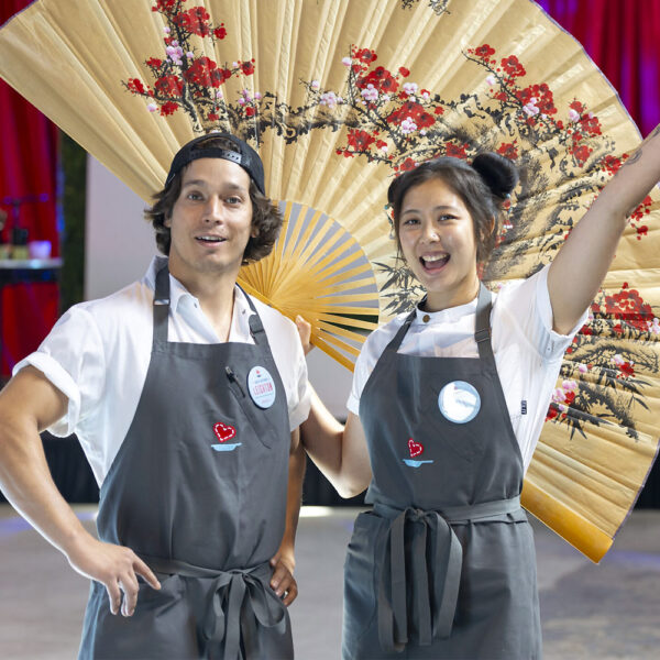 Chefs Sarah Cai with her right arm in the air and Chef Arturo Leighton smiling for the camera in front of a large foldable paper fan decoration